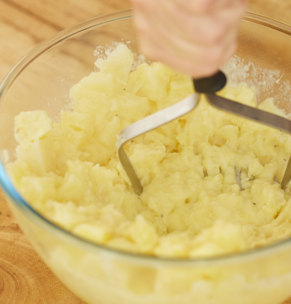 A close-up view of cooked yellow potatoes being mashed with a stainless steel hand masher in a clear glass bowl on a wooden surface.
