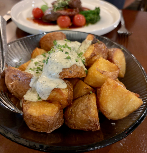 A bowl of golden crispy four-times cooked roast potatoes topped with creamy caper and herb mayonnaise at Sandalford Winery, with a main dish in the background.