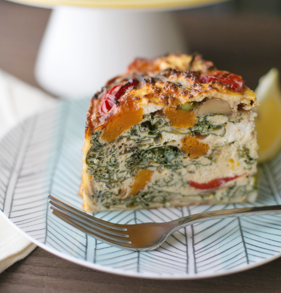 A thick slice of baked gluten-free potato frittata layered with spinach, pumpkin, and cherry tomatoes, served on a patterned plate with a silver fork.