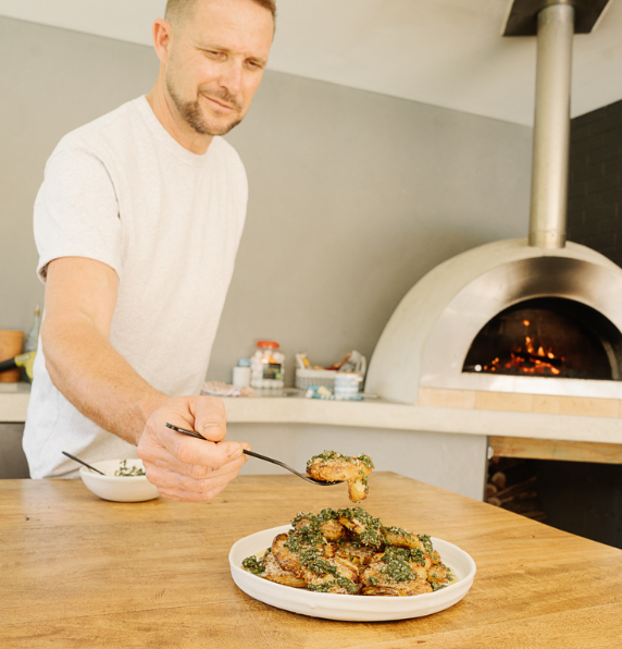 Chef Scott Bridger using tongs to plate crispy smash roasted potatoes topped with zesty green chimichurri sauce, with a wood-fired oven in the background.