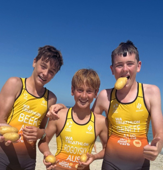 Three young triathletes in yellow and orange WA Triathlon uniforms smiling and playfully posing on a beach while holding raw potatoes.