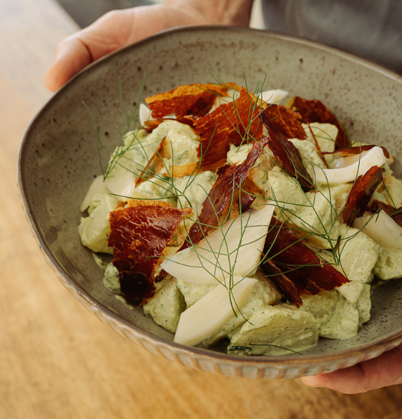 A close-up view of hands holding a gray textured bowl filled with creamy herb potato salad, topped with shards of crispy prosciutto and fresh dill.